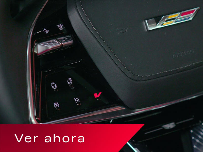 Vista en primer plano del volante de una Cadillac OPTIQ, en la que se exhiben los controles táctiles iluminados y un prominente emblema de V-Series arriba de un cartel rojo en el que se lee "Watch Now" (Mirar ahora).