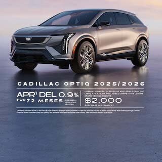 Imagen de un SUV eléctrico Cadillac OPTIQ plateado estacionado sobre una superficie lisa y reflectante al anochecer, visto desde una perspectiva frontal de tres cuartos. El texto grande dice "Modelos Cadillac OPTIQ 2025/2026", "0.9% APR durante 72 meses," y "$2,000 de descuento en la compra," con detalles de la oferta en letra pequeña en la parte inferior.